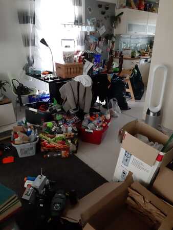 Unordentliches Wohnzimmer mit vielen Kartons, Verpackungen und Haushaltsgegenständen im Chaos.