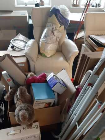 Wohnzimmer mit Umzugskartons, Sessel und Haushaltsgegenständen in chaotischer Anordnung.